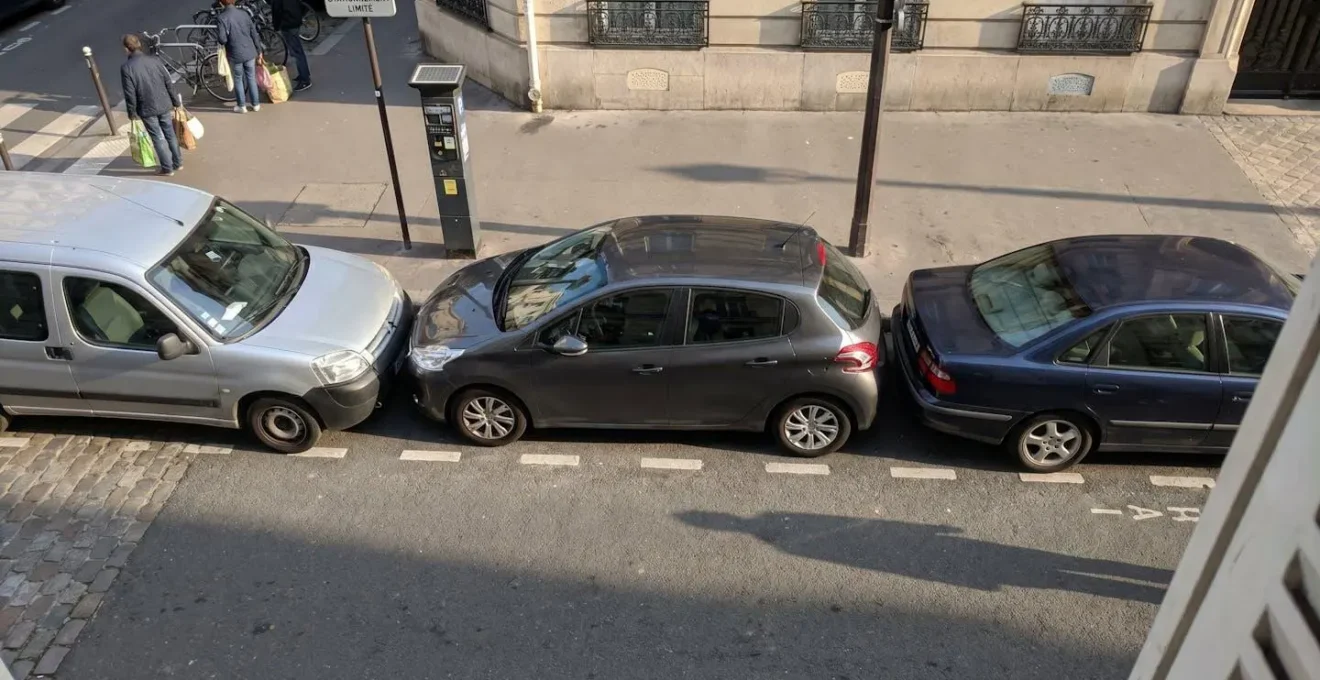 Petite citadine blanche garée en créneau dans un espace de stationnement urbain très serré, vue en plongée montrant les faibles distances avec les véhicules adjacents sur une rue pavée